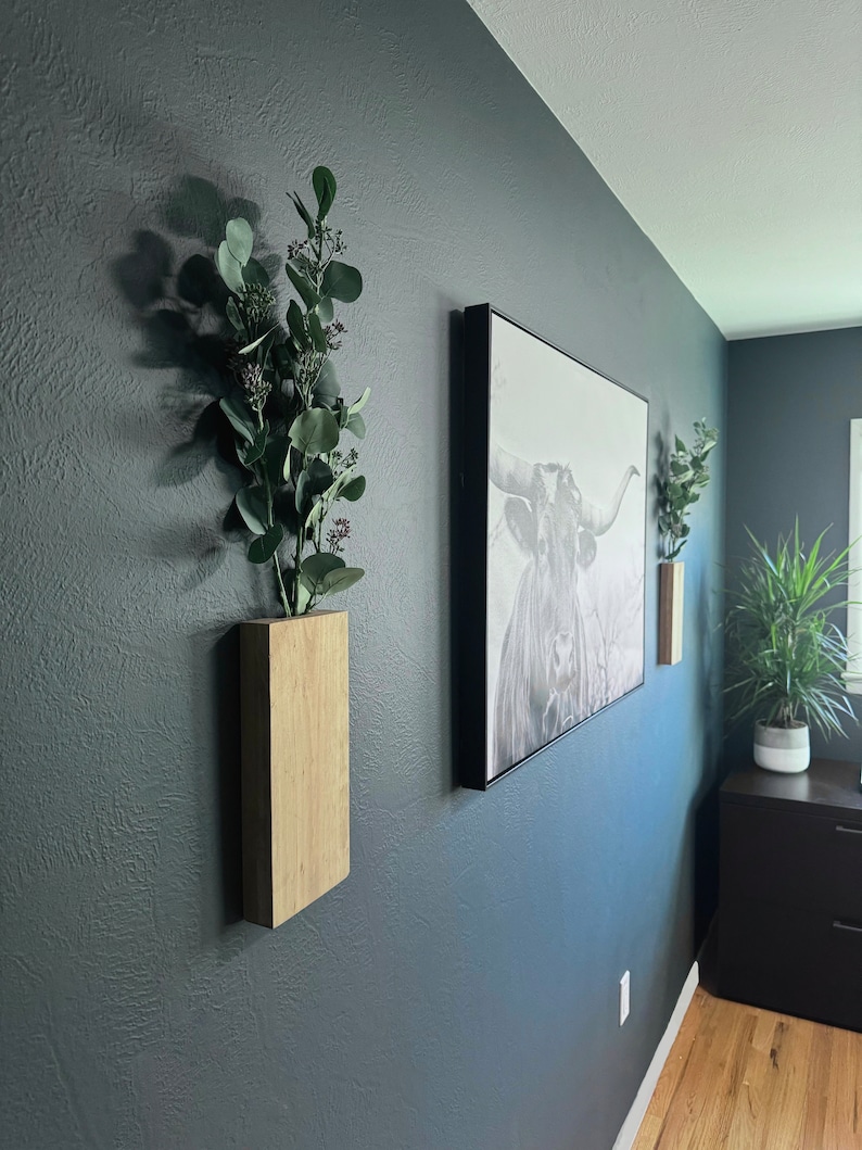 Op de afbeelding: Twee houten wandplanters met groen in een donkerblauwe kamer. Een zwart ingelijste print van een stier hangt aan de muur boven de planters.