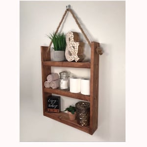 May include: A rustic wooden shelf with three tiers, hanging from a rope and a hook. The shelf is decorated with a small plant, a decorative ampersand, towels, a glass jar, toilet paper rolls, a small plant, and a candle holder. The shelf is stained a dark brown color.