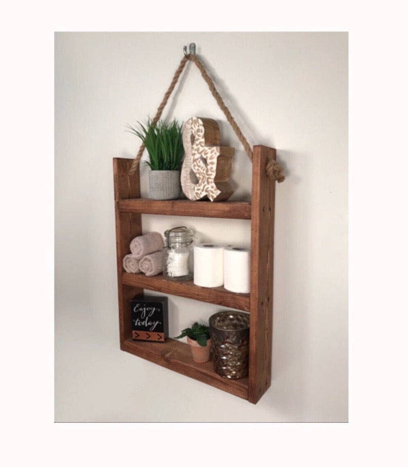 May include: A rustic wooden shelf with three tiers, hanging from a rope and a hook. The shelf is decorated with a small plant, a decorative ampersand, towels, a glass jar, toilet paper rolls, a small plant, and a candle holder. The shelf is stained a dark brown color.