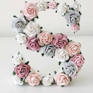 K&ouml;nnte beinhalten: Ein wei&szlig;er Buchstabe S, der mit rosa, wei&szlig;en und grauen Papierblumen bedeckt ist. Die Blumen sind in einem Kreis um den Buchstaben angeordnet.