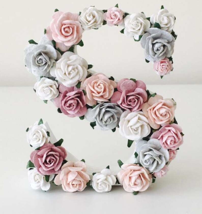 K&ouml;nnte beinhalten: Ein wei&szlig;er Buchstabe S, der mit rosa, wei&szlig;en und grauen Papierblumen bedeckt ist. Die Blumen sind in einem Kreis um den Buchstaben angeordnet.