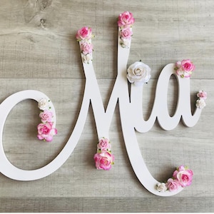 May include: A white wooden sign spelling "Mia" in cursive, decorated with pink and white artificial flowers. The sign is set against a light wood-grain background. The flowers are in shades of pink and white.