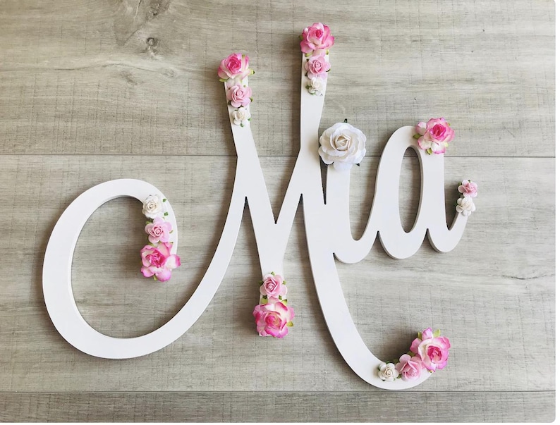 May include: A white wooden sign spelling "Mia" in cursive, decorated with pink and white artificial flowers. The sign is set against a light wood-grain background. The flowers are in shades of pink and white.