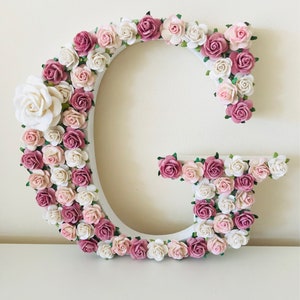 Pode incluir: Uma letra G de madeira branca decorada com flores de papel rosa e branco. As flores est&atilde;o dispostas em um padr&atilde;o circular ao redor da letra.