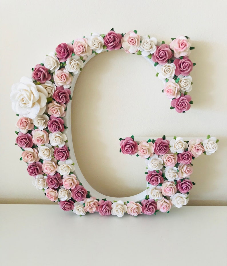 Pode incluir: Uma letra G de madeira branca decorada com flores de papel rosa e branco. As flores est&atilde;o dispostas em um padr&atilde;o circular ao redor da letra.