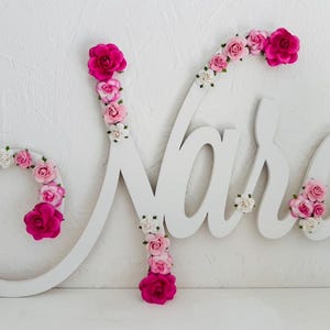 May include: White wooden name sign spelling "Nara" in cursive, decorated with pink and white flowers. The letter "N" and "a" are adorned with vibrant pink roses. A decorative piece for a nursery or bedroom.