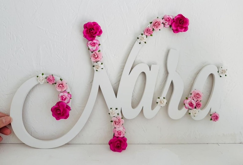 May include: White wooden name sign spelling "Nara" in cursive, decorated with pink and white flowers. The letter "N" and "a" are adorned with vibrant pink roses. A decorative piece for a nursery or bedroom.