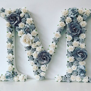 K&ouml;nnte beinhalten: Ein wei&szlig;er Buchstabe M, der mit wei&szlig;en und grauen Papierblumen bedeckt ist. Die Blumen sind in einem zuf&auml;lligen Muster angeordnet und erzeugen einen 3D-Effekt.