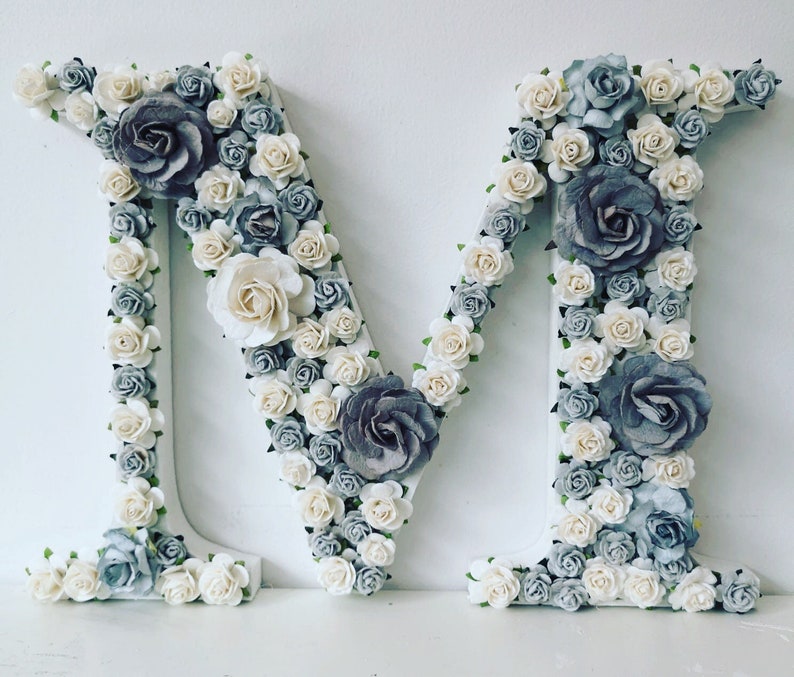 K&ouml;nnte beinhalten: Ein wei&szlig;er Buchstabe M, der mit wei&szlig;en und grauen Papierblumen bedeckt ist. Die Blumen sind in einem zuf&auml;lligen Muster angeordnet und erzeugen einen 3D-Effekt.