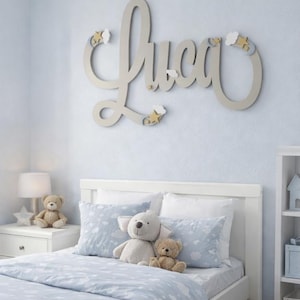 May include: A nursery scene with a white bed, blue bedding, and stuffed animals. A wall decoration spells out the name "Luca" in cursive with star and cloud accents. A nightstand with a lamp and teddy bear is on the left.