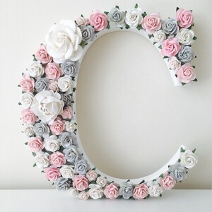 Pode incluir: Uma letra C de madeira branca decorada com flores de papel rosa, brancas e cinzas. As flores est&atilde;o dispostas em um padr&atilde;o circular ao redor da letra.