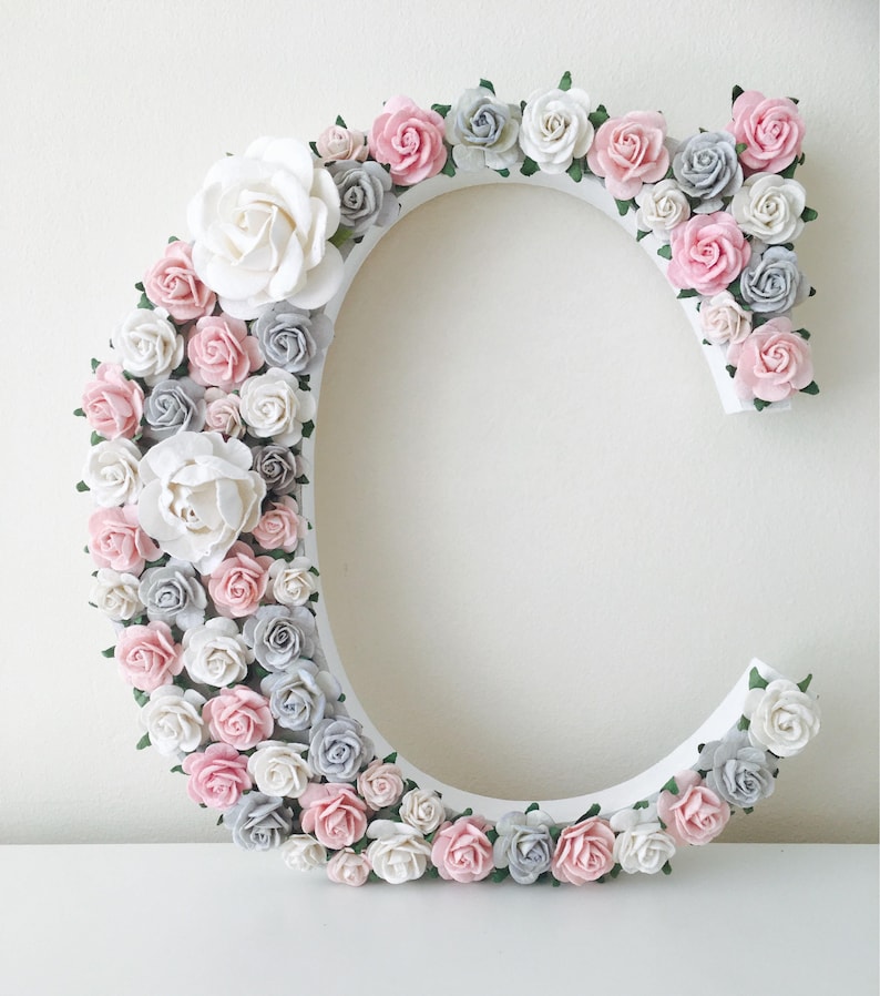 Pode incluir: Uma letra C de madeira branca decorada com flores de papel rosa, brancas e cinzas. As flores est&atilde;o dispostas em um padr&atilde;o circular ao redor da letra.
