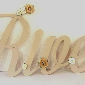 May include: A wooden sign spelling "Rueen" in a cursive font. The letters are a light beige colour and decorated with small, cream and tan paper roses. The sign is designed for wall decor, suitable for a nursery.