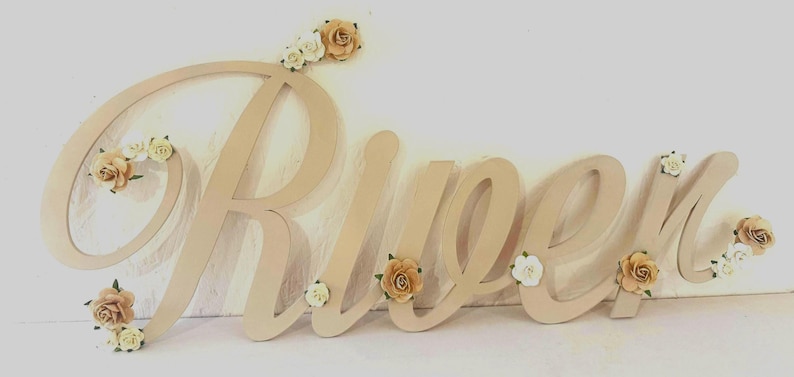 May include: A wooden sign spelling "Rueen" in a cursive font. The letters are a light beige colour and decorated with small, cream and tan paper roses. The sign is designed for wall decor, suitable for a nursery.