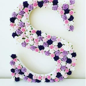 K&ouml;nnte beinhalten: Ein wei&szlig;er Buchstabe S, der mit kleinen k&uuml;nstlichen Blumen in Rosa-, Lila- und Wei&szlig;t&ouml;nen bedeckt ist.