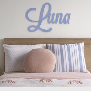 May include: A pale blue wooden sign spelling "Luna" is above a bed with a wooden headboard. The bed has patterned pillows and a pink comforter with rainbow designs. A pink alarm clock and lamp are on the bedside table.