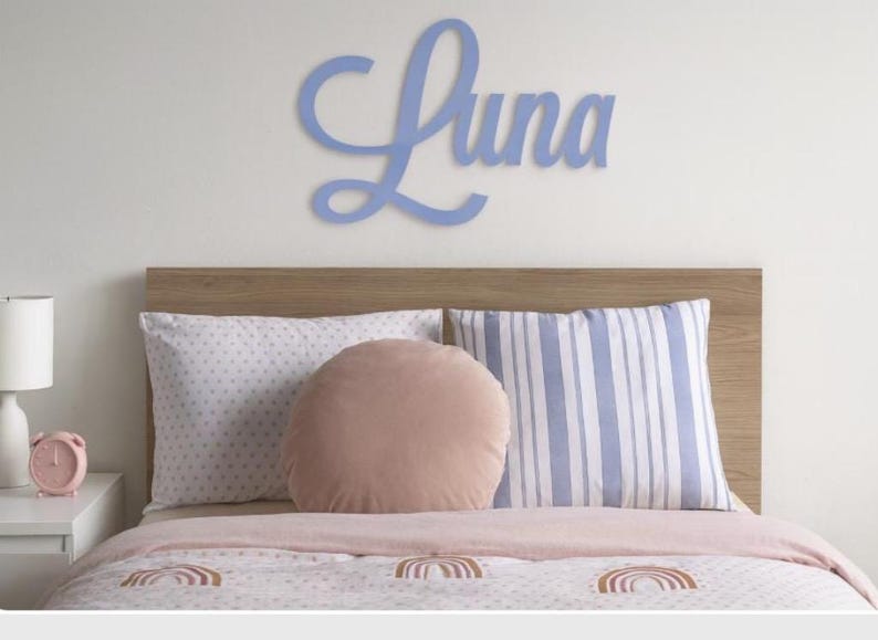 May include: A pale blue wooden sign spelling "Luna" is above a bed with a wooden headboard. The bed has patterned pillows and a pink comforter with rainbow designs. A pink alarm clock and lamp are on the bedside table.