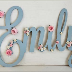 May include: A light blue wooden name sign spelling "Emily", decorated with pink and white paper flowers. The letters are in a script font and are displayed on a white surface.