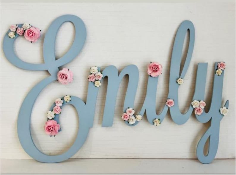 May include: A light blue wooden name sign spelling "Emily", decorated with pink and white paper flowers. The letters are in a script font and are displayed on a white surface.