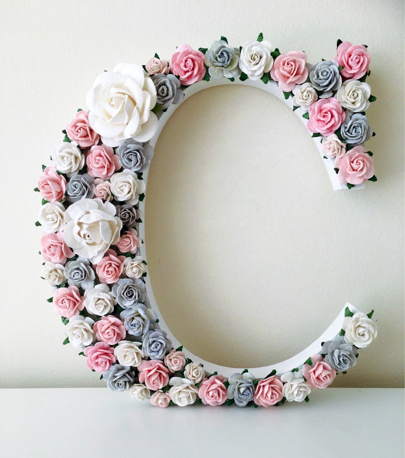K&ouml;nnte beinhalten: Ein wei&szlig;er Buchstabe C, der mit rosa, grauen und wei&szlig;en Papierblumen verziert ist. Der Buchstabe ist aus Holz und eine tolle Erg&auml;nzung f&uuml;r ein Kinderzimmer oder ein Babyzimmer.
