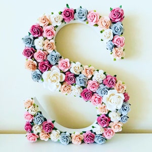 K&ouml;nnte beinhalten: Ein wei&szlig;er Holzbuchstabe S, der mit rosa, blauen und cremefarbenen Papierblumen verziert ist. Die Blumen sind in einem Kreis um den Buchstaben angeordnet.