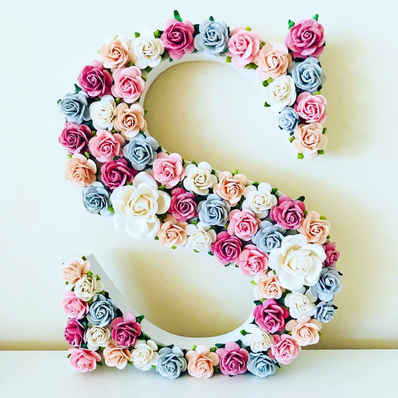 K&ouml;nnte beinhalten: Ein wei&szlig;er Holzbuchstabe S, der mit rosa, blauen und cremefarbenen Papierblumen verziert ist. Die Blumen sind in einem Kreis um den Buchstaben angeordnet.