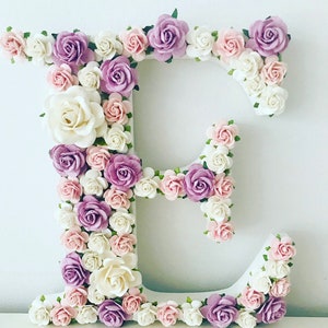 Pode incluir: Uma letra E branca decorada com flores de papel rosa, brancas e roxas. As flores est&atilde;o dispostas em um padr&atilde;o circular ao redor da letra.