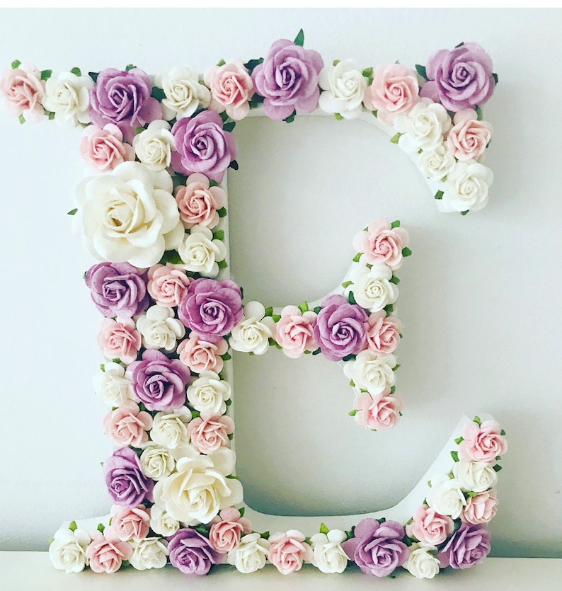 Pode incluir: Uma letra E branca decorada com flores de papel rosa, brancas e roxas. As flores est&atilde;o dispostas em um padr&atilde;o circular ao redor da letra.