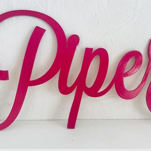 May include: A magenta wooden sign spelling out "Piper" in a cursive font. The sign is painted in a bright pink hue. Ideal for decorating a child's bedroom or nursery, this sign adds a personal touch.