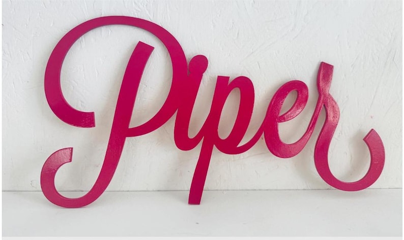 May include: A magenta wooden sign spelling out "Piper" in a cursive font. The sign is painted in a bright pink hue. Ideal for decorating a child's bedroom or nursery, this sign adds a personal touch.