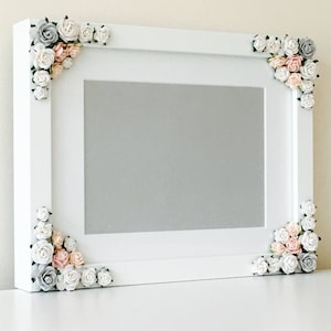 Pode incluir: Uma moldura retangular branca com interior cinza. Os cantos são adornados com aglomerados de pequenas rosas artificiais em tons de branco, rosa e cinza. A moldura é exibida em uma superfície branca.