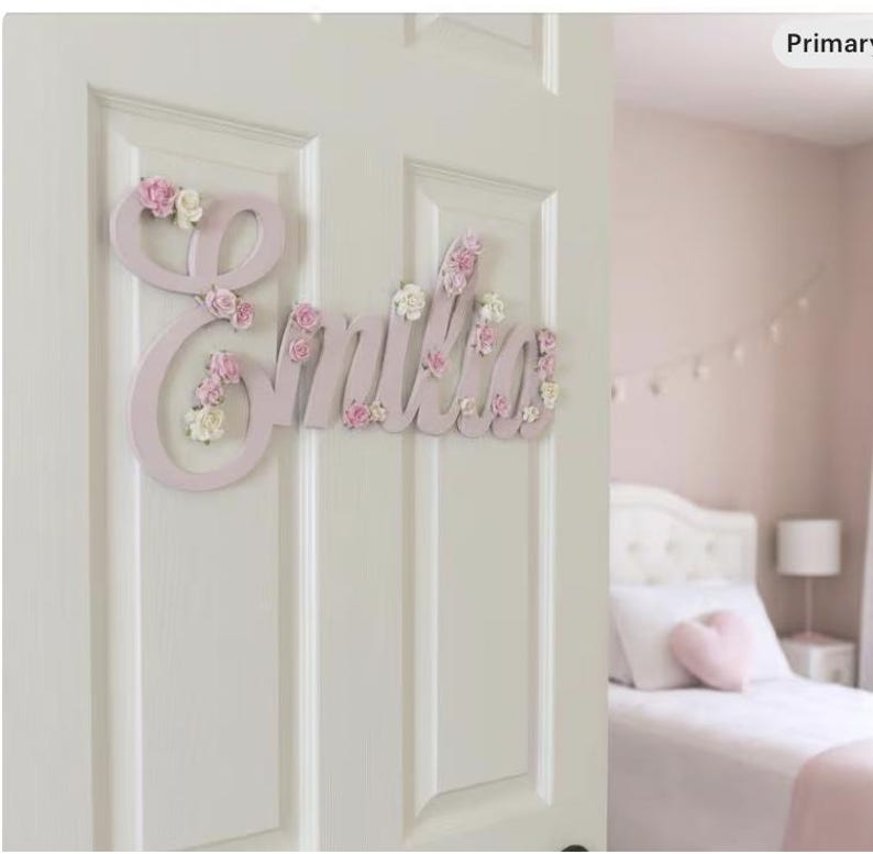 May include: A pale pink wooden name sign spelling "Emilia" in a cursive font, decorated with small pink and white flowers. The sign is fixed to a white door, suitable for a child's bedroom.