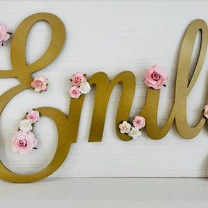 May include: A gold-coloured wooden name sign spelling "Emily" with pink and white floral decorations. The cursive letters are ideal for nursery decor or personalised gifts.