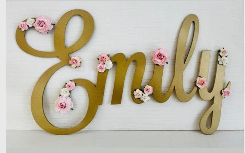 May include: A gold-coloured wooden name sign spelling "Emily" with pink and white floral decorations. The cursive letters are ideal for nursery decor or personalised gifts.