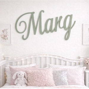 May include: A child's bedroom with a white cot and pink bedding, including cushions and a stuffed bunny. A large, cursive "Marg" sign in green is on the wall, alongside framed bunny and floral artwork.