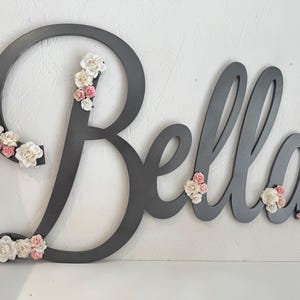 May include: A grey wooden sign spelling "Bella" with floral accents. The cursive letters are decorated with small, white and pink flowers. The sign is held by a hand against a white background.