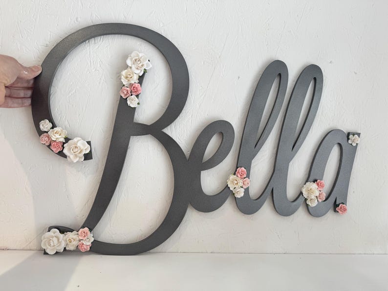 May include: A grey wooden sign spelling "Bella" with floral accents. The cursive letters are decorated with small, white and pink flowers. The sign is held by a hand against a white background.
