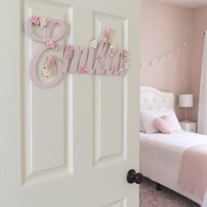 May include: A light pink wooden name sign, "Emilia", is mounted on a white door. The letters are decorated with small pink and white floral accents. The bedroom in the background has a pink colour scheme.