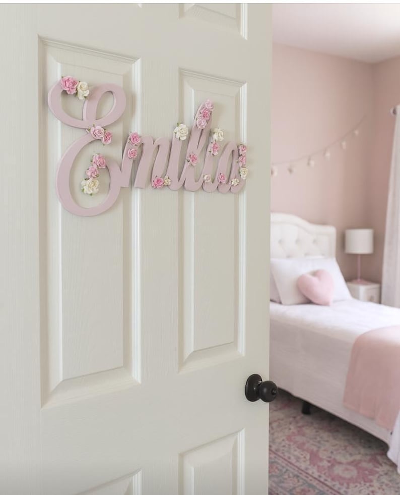May include: A light pink wooden name sign, "Emilia", is mounted on a white door. The letters are decorated with small pink and white floral accents. The bedroom in the background has a pink colour scheme.