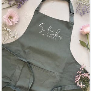 May include: A sage green apron featuring an adjustable neck strap and a front pocket. The chest area displays the white printed text "She cooks BY CHLOE" in a combination of cursive and block lettering, centred on the fabric surface.