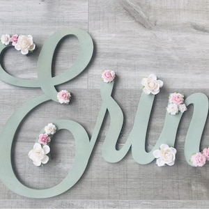 May include: A wooden sign spelling out the name "Erin" in a soft green hue. The letters are decorated with delicate, handcrafted white and pink paper flowers. This sign is ideal for nursery decor, adding a touch of elegance.