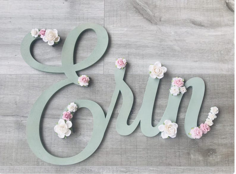 May include: A wooden sign spelling out the name "Erin" in a soft green hue. The letters are decorated with delicate, handcrafted white and pink paper flowers. This sign is ideal for nursery decor, adding a touch of elegance.