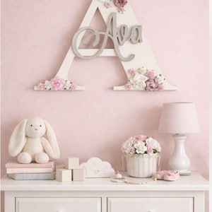 Puede incluir: Una letra "A" de madera blanca decorada con flores rosas y blancas, con el nombre "Ava" en plata. La letra está montada en una pared rosa sobre una cómoda blanca con un conejito de peluche y una lámpara.
