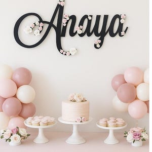 May include: A party scene with a pink theme. A black cursive "Anaya" sign is decorated with flowers. A cake and cupcakes sit on white stands. Balloons in shades of pink and white are arranged on either side of the table. Flowers in white vases complete the decor.