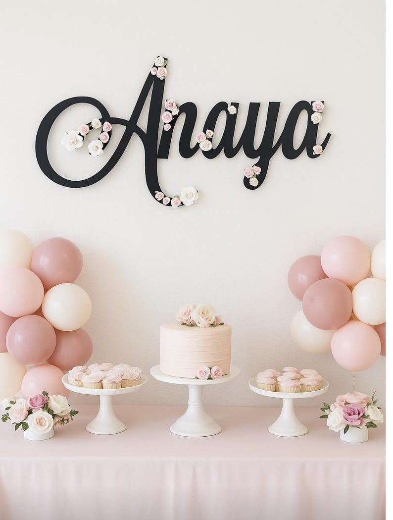 May include: A party scene with a pink theme. A black cursive "Anaya" sign is decorated with flowers. A cake and cupcakes sit on white stands. Balloons in shades of pink and white are arranged on either side of the table. Flowers in white vases complete the decor.
