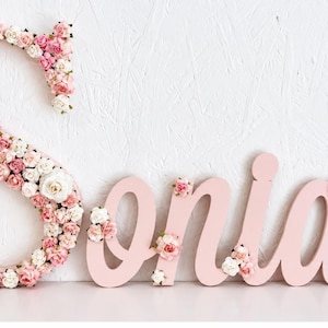 Könnte beinhalten: Ein dekoratives Wandschild, das den Namen Soniah in einer Schreibschrift buchstabiert. Der Buchstabe S ist mit kleinen künstlichen rosa und weißen Rosen bedeckt, während die Buchstaben oniah hellrosa lackiert und mit einzelnen Rosen verziert sind.