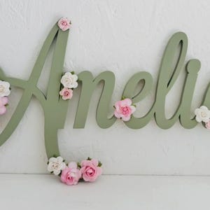 May include: A sage green wooden name sign spelling "Amelia" in a flowing script. The letters are decorated with clusters of small pink and white paper flowers. Ideal for a child's bedroom or nursery decoration.