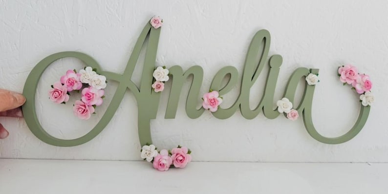 May include: A sage green wooden name sign spelling "Amelia" in a flowing script. The letters are decorated with clusters of small pink and white paper flowers. Ideal for a child's bedroom or nursery decoration.