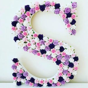 Pode incluir: Uma letra S branca coberta de pequenas flores artificiais em tons de rosa, roxo e branco.