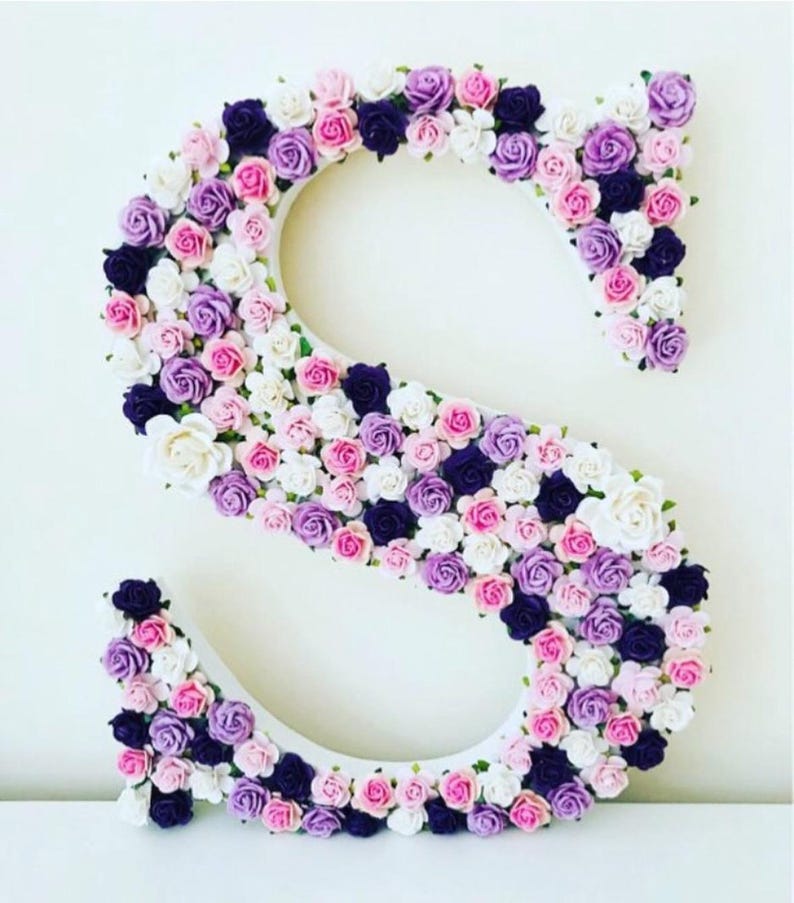 Pode incluir: Uma letra S branca coberta de pequenas flores artificiais em tons de rosa, roxo e branco.